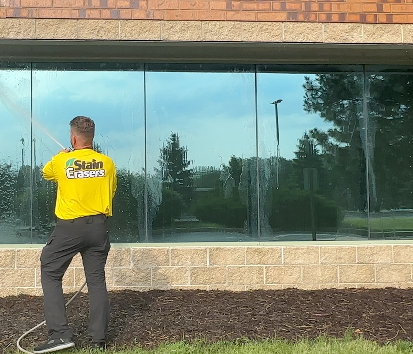A Stain Erasers technician soft-washing a wall of storefront windows on a bright day